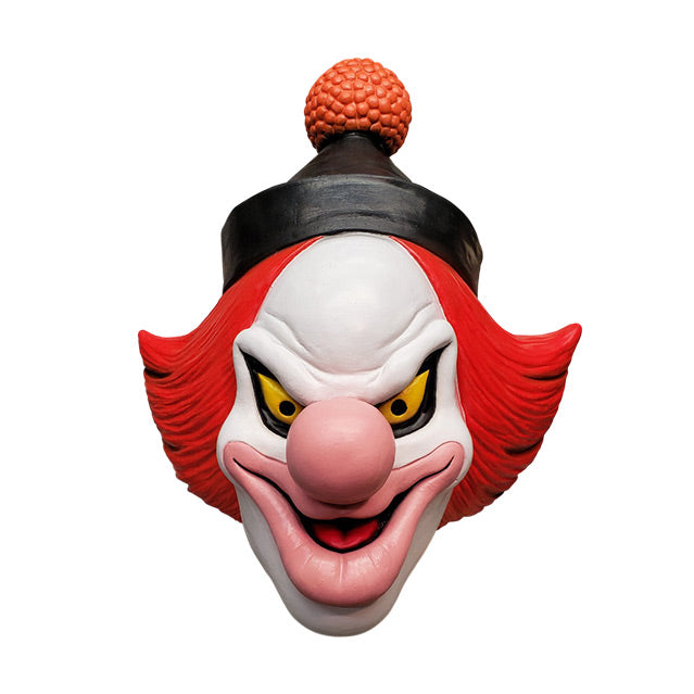 Mask, front view.  White clown face, Black-rimmed upturned yellow eyes, large pale pink nose and lips, menacing smile with mouth slightly open, showing red tongue.  Red hair, black cap with orange pompom at top.