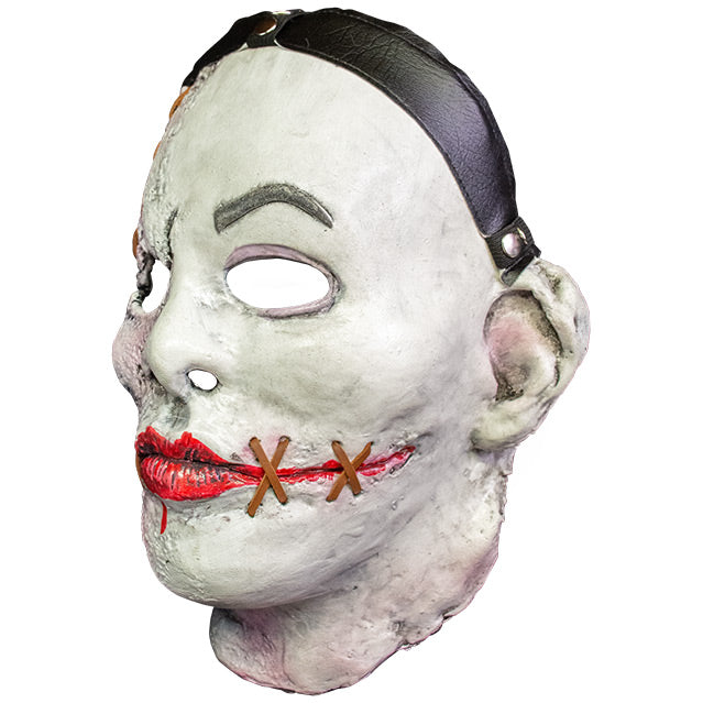 Mask, head and neck, left side view. White flesh, black strap across hairline, distressed skin on right side of face, brown stiches on forehead above right eye and on sides of painted on red mouth. Gray left eyebrow.
