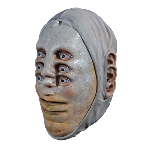 Mask. Left side view. Pale white skin. 3 sets of pale blue eyes one above the other, 3 noses one above the other in center of face, two mouths, one above the other at bottom of face, 3 ears on each side of head. White cloth wrap around head to edges of face.