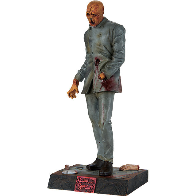 Statue, left side view. Zombie figure, orange hued decaying flesh, wearing distressed, dirty, bloodied blue doctor's coat and blue pants, black shoes, missing left hand. Standing on black, dirty base, severed hand and hatchet are resting on base, plaque on front red text reads house by the Cemetary.