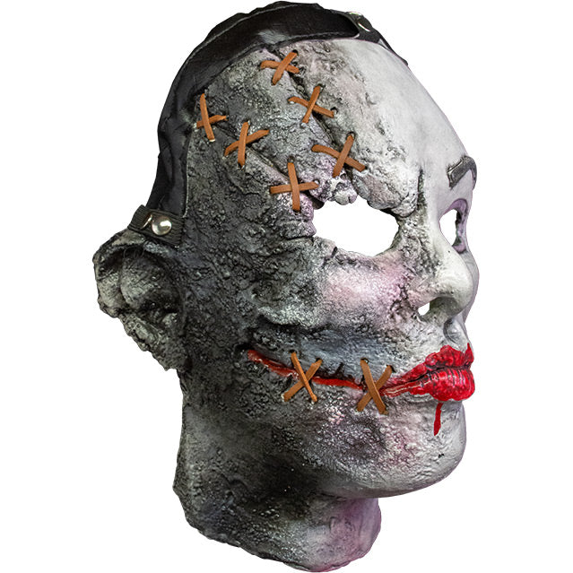 Mask, head and neck, right side view. White flesh, black strap across hairline, distressed skin on right side of face, brown stiches on forehead above right eye and on sides of painted on red mouth. Gray left eyebrow.