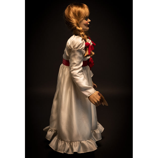 Doll, right side view, black background. Blond hair with bangs, two braids tied with red ribbon. White, floor-length dress with red trim at chest and red belt with rose at waist. Holding card in right hand.