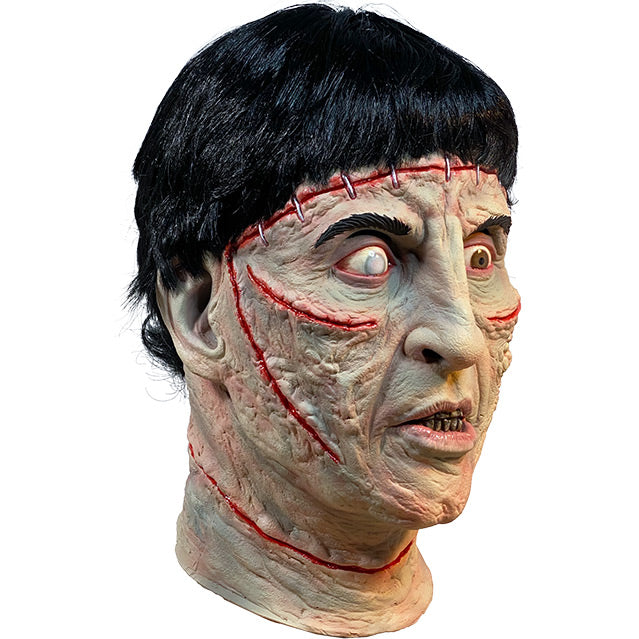 Mask, right side view, head and neck. Short black hair, staples in forehead, red wounds below eyes, on cheeks and on neck, red-rimmed eyes, right eye iris is white, left eye iris is brown. Mouth slightly open, showing teeth.