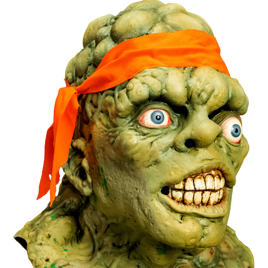 Mask, head, neck and upper chest. Right, close up view. Green lumpy blistered flesh, bald with bright orange headband tied around forehead. Misaligned blue eyes, crooked nose. Lips open showing large dirty teeth. Bumpy neck and chest with sores.