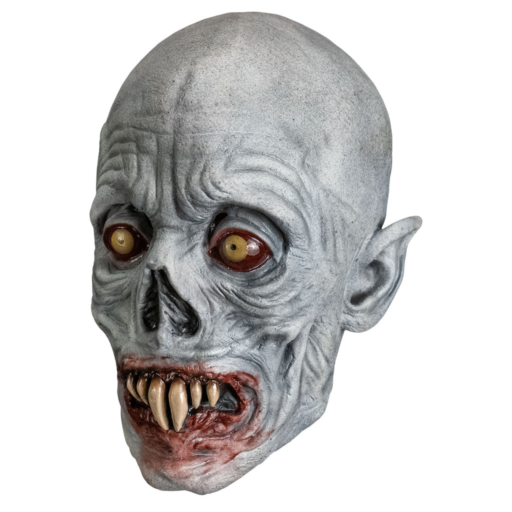 Mask, left view. Bald, gray wrinkled skin, no eyebrows, sunken yellow and red eyes, pointed ears, skeletal nose, blood-rimmed grimacing mouth, two long central fangs with two smaller fangs on either side.