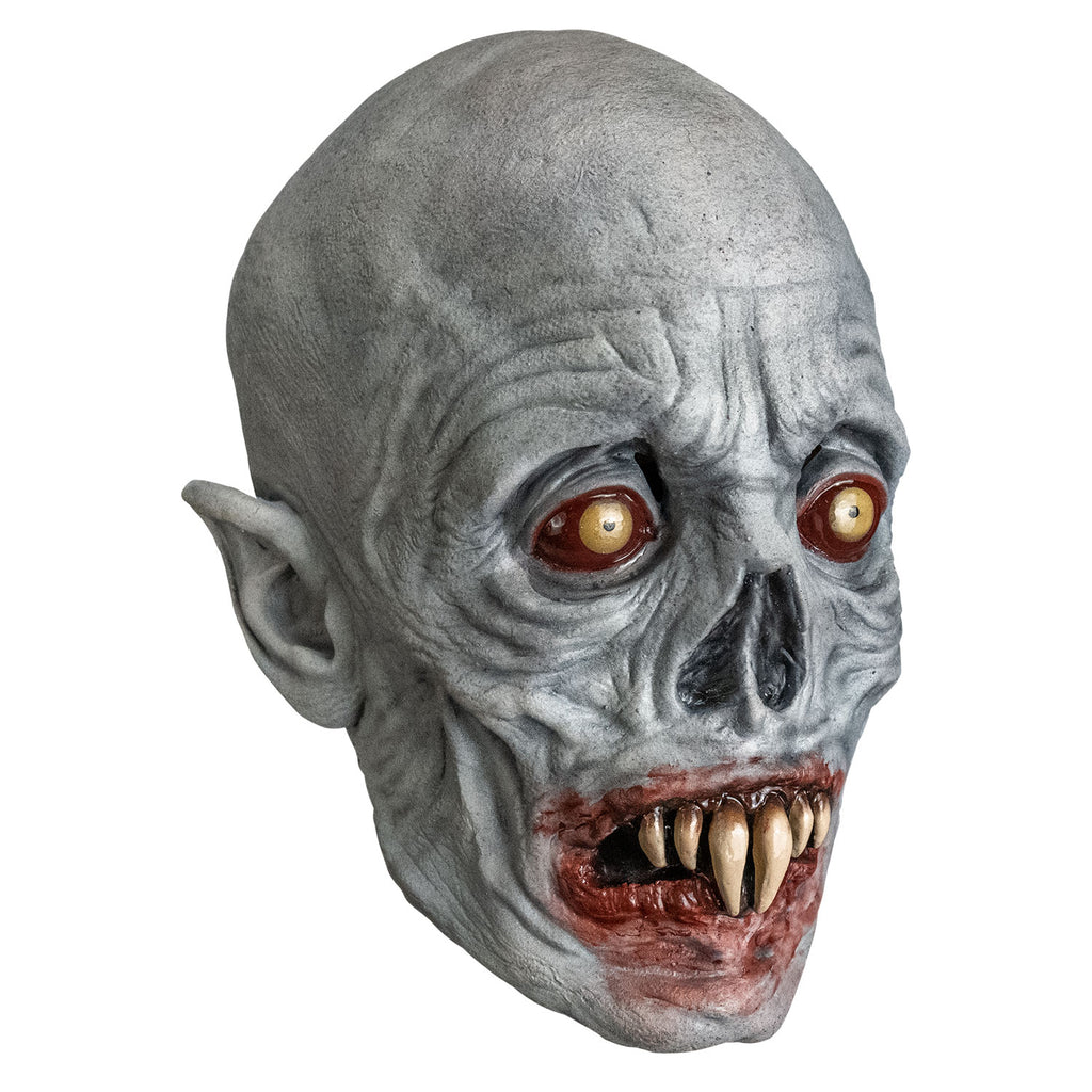 Mask, right view. Bald, gray wrinkled skin, no eyebrows, sunken yellow and red eyes, pointed ears, skeletal nose, blood-rimmed grimacing mouth, two long central fangs with two smaller fangs on either side.