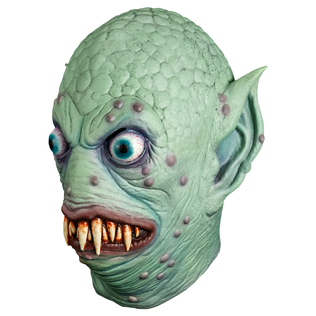 Mask, left side view. Pale blue-green scaly flesh on top of head, wrinkled around eyes, mouth and neck, scattered pale purple bumps on face and ears. Large pointed ears. Bulging, bloodshot, round aqua blue eyes. No nose. pale red, lumpy lips slightly open showing may sharp dirty brown teeth.