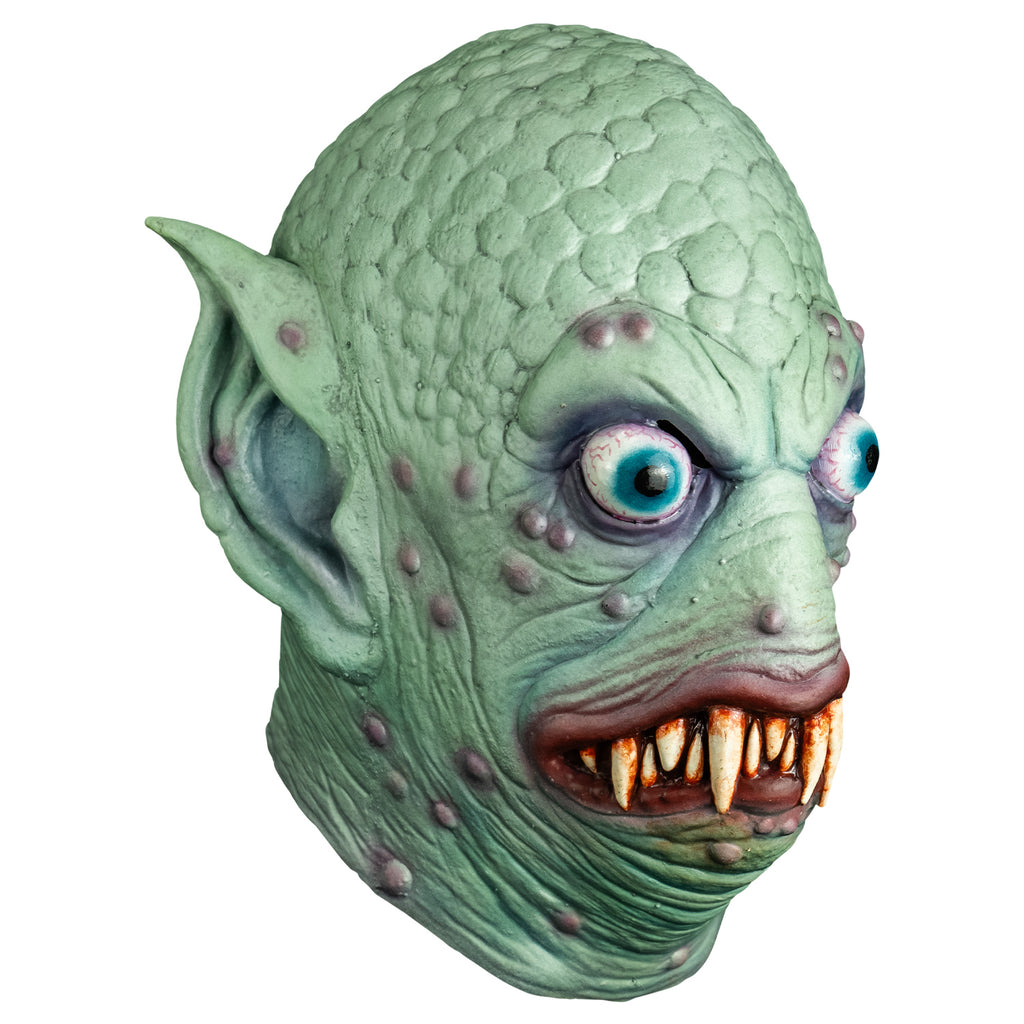 Mask, right side view. Pale blue-green scaly flesh on top of head, wrinkled around eyes, mouth and neck, scattered pale purple bumps on face and ears. Large pointed ears. Bulging, bloodshot, round aqua blue eyes. No nose. pale red, lumpy lips slightly open showing may sharp dirty brown teeth.