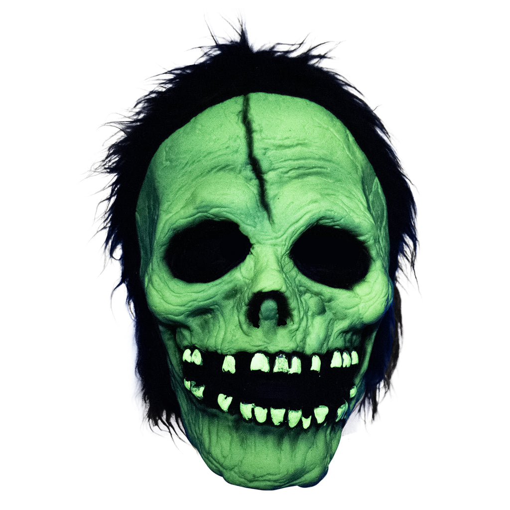 Mask, front view. Green glow feature shown skull-like face. Black, short shaggy hair, black crack down the forehead. Black rimmed empty eyes, skull like nasal cavity. black, open grinning mouth with several missing teeth