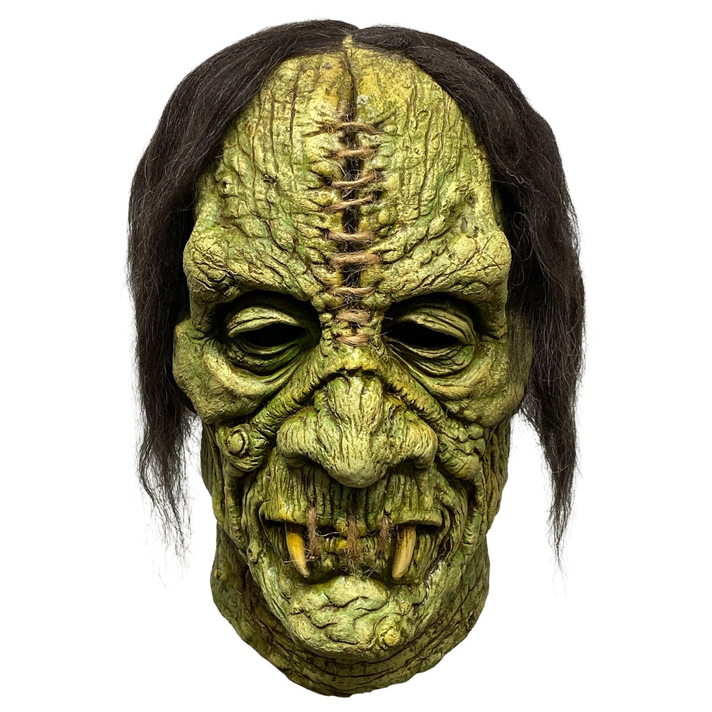 Mask, front view.  Stringy black hair.  Greenish wrinkled skin with warts. Large gash in center of forehead, stitched with brown twine.  Mouth stitched closed with brown twine, two long yellow fangs.
