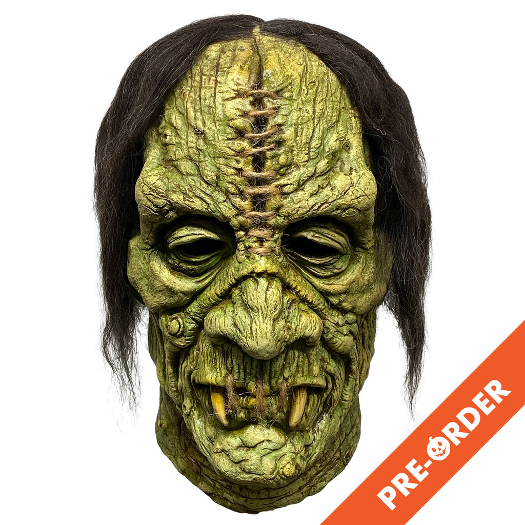 white background, orange diagonal banner bottom right, white text reads pre-order.  Mask, front view. Stringy black hair. Greenish wrinkled skin with warts. Large gash in center of forehead, stitched with brown twine. Mouth stitched closed with brown twine, two long yellow fangs.