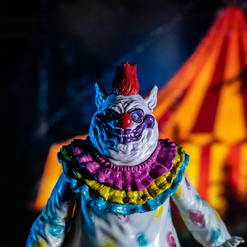 glamour shot close up.  nighttime circus background.  Close up of Fatso Figure, front view. Lumpy and wrinkly face with several flesh folds beneath chin, large pointed ears on side of head. White skin, red hair on top, purple around eyes, large dark pink nose and lumpy purple clown mouth. wearing white clown suit with pink, blue and yellow circles on it. Pink, yellow and blue ruffles at neck.