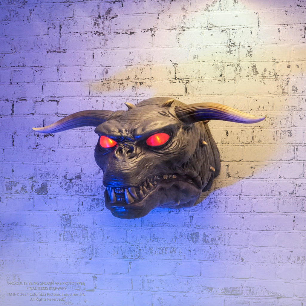 white painted brick wall background. Wall mounted demon dog head. Gray wrinkled skin, short square muzzle with sharp teeth, small pug-like nose between lighted red eyes.  two large horns on either side of head smaller spikes on top and sides.