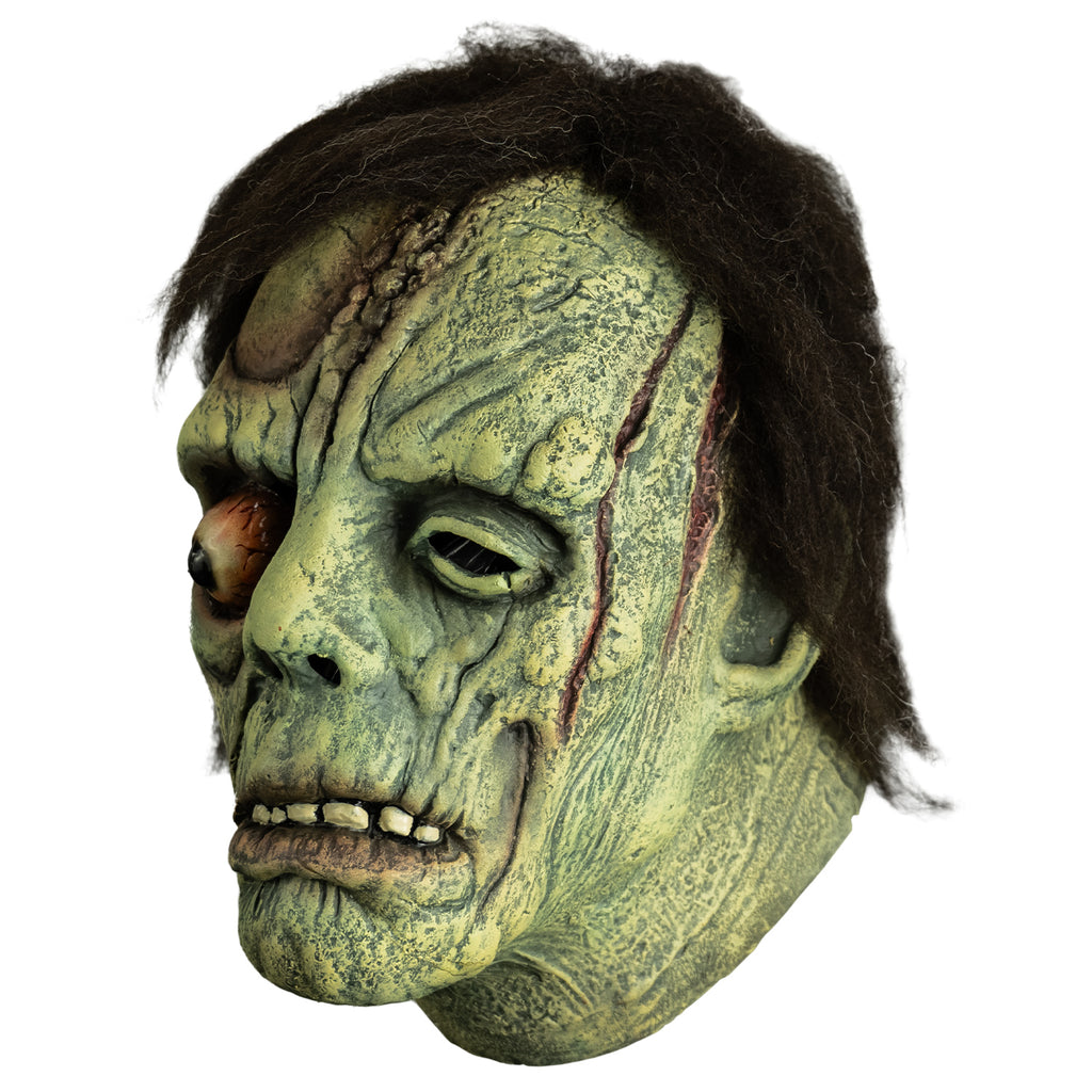 Mask, left side view. Short black hair. Greenish flesh, wrinkled and scarred. Bulging right eye. Mouth, slightly open showing bottom teeth, prominent lower lip