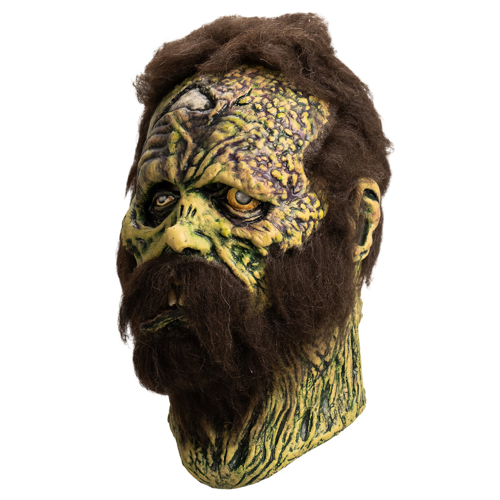 Mask, left side view. Open wound on right forehead revealing skull underneath. short, matted brown hair, bushy brown sideburns and full beard with mustache. no eyebrows. Mottled, wrinkled and sagging yellow-green and brown skin. Right eye squinting almost shut. Bloodshot, open yellow left eye. small deformed nose. Crooked mouth, slightly open showing large front teeth