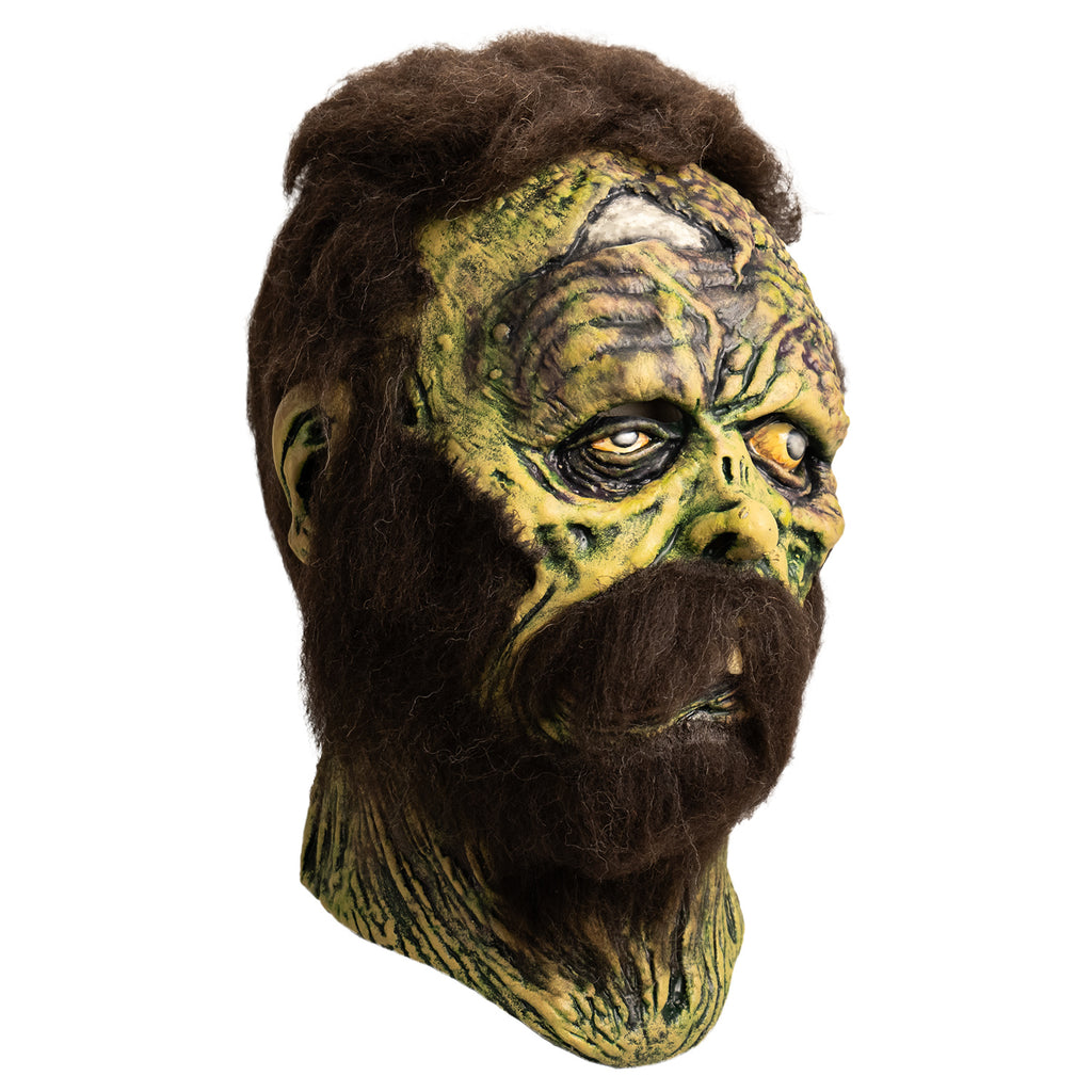 Mask, right side view. Open wound on right forehead revealing skull underneath. short, matted brown hair, bushy brown sideburns and full beard with mustache. no eyebrows. Mottled, wrinkled and sagging yellow-green and brown skin. Right eye squinting almost shut. Bloodshot, open yellow left eye. small deformed nose. Crooked mouth, slightly open showing large front teeth
