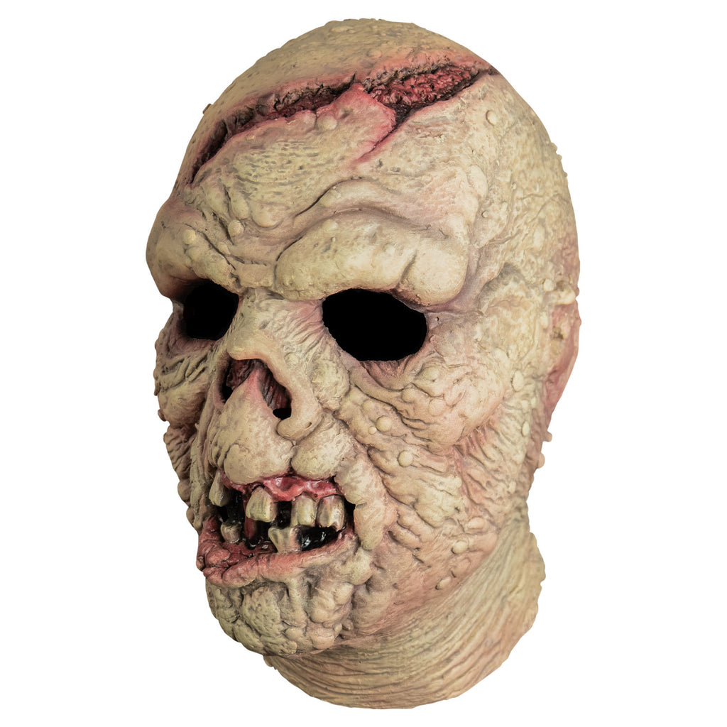 Mask, left side view, head and neck. Bald. Pinkish, wrinkled and lumpy flesh, large open wound on forehead and crown of head. missing nose. Mouth open showing gums, gapped dirty teeth, deformed pink bottom lip.