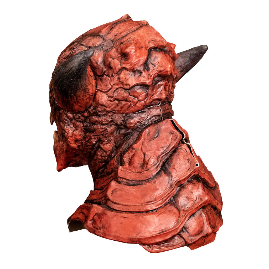 Mask, back left side view. Red creature with horns on either side of head, wrinkled and scaly skin on face pointed chin. Large plate-like scales on back of head, neck and chest