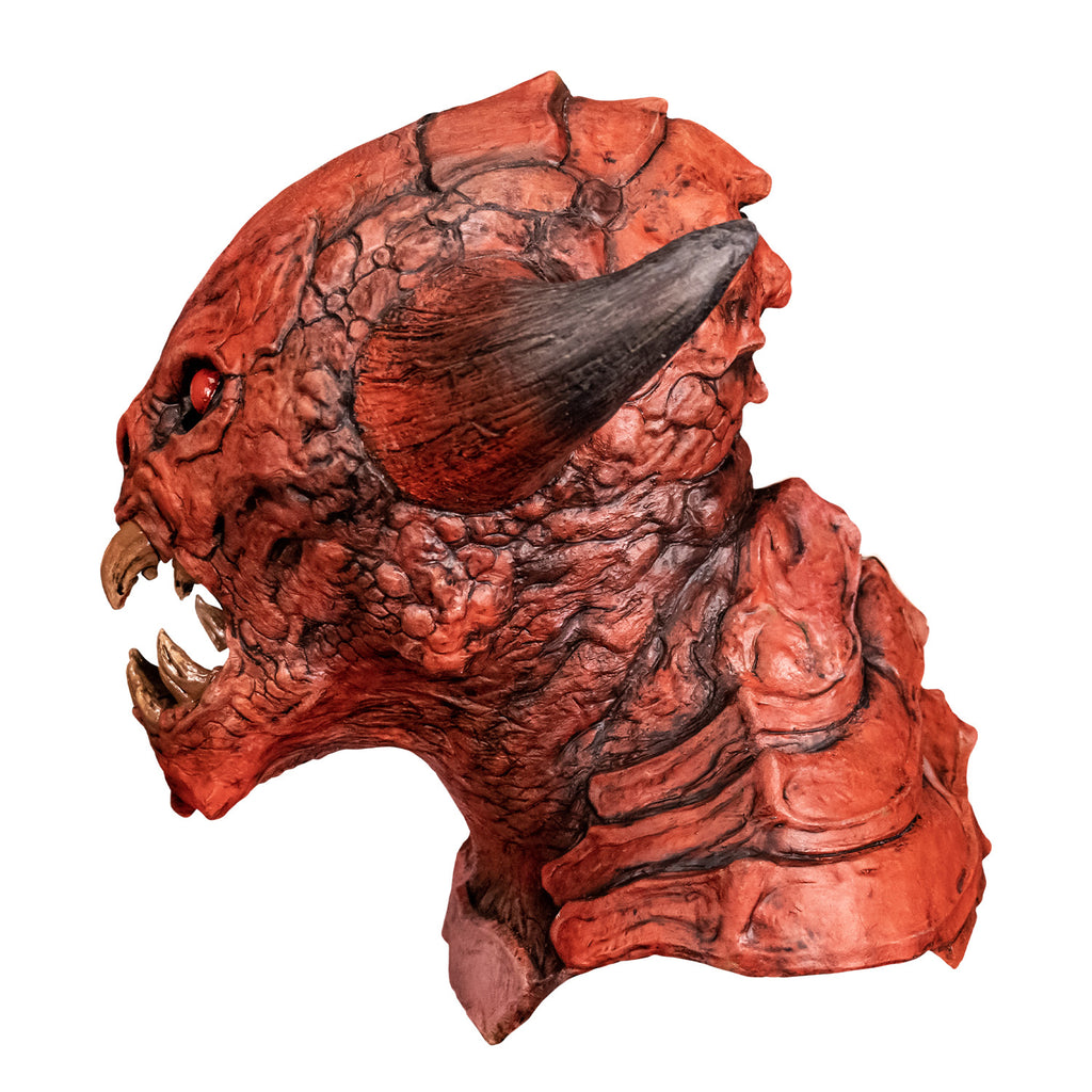 Mask, left profile view. Red creature with horns on either side of head, red black-rimmed eyes, snout-like nose, open mouth showing several large yellowed sharp fangs, wrinkled and scaly skin on face pointed chin. Large plate-like scales on back of head, neck and chest