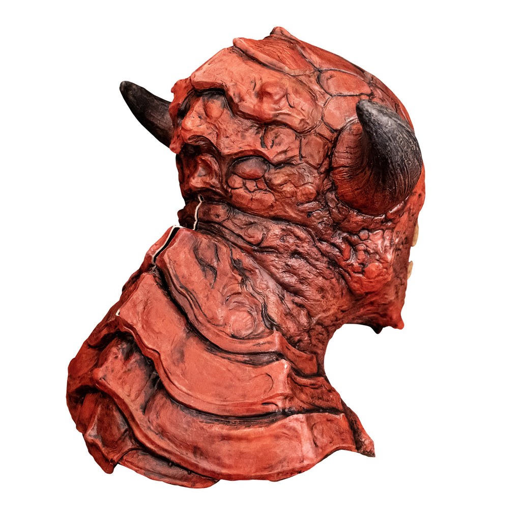 Mask, back right side view. Red creature with horns on either side of head,  wrinkled and scaly skin on face pointed chin. Large plate-like scales on back of head, neck and chest