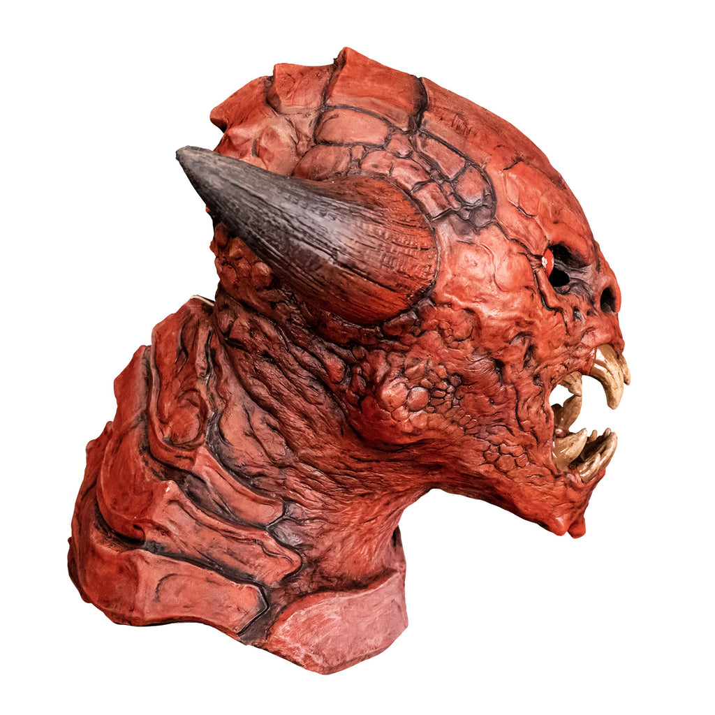 Mask, right profile view. Red creature with horns on either side of head, red black-rimmed eyes, snout-like nose, open mouth showing several large yellowed sharp fangs, wrinkled and scaly skin on face pointed chin. Large plate-like scales on back of head, neck and chest