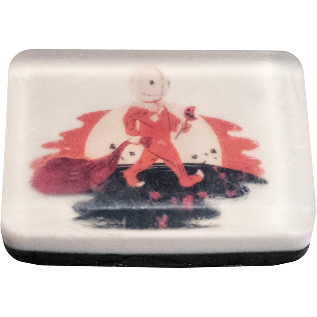 Bar soap. White, with illustration under clear soap layer of Trick r' Treat Sam, sewn burlap mask, orange jumpsuit, holding bag in right hand, bitten jack o' lantern lollipop in left hand, orange and white moon background, black and orange middle and foreground. 