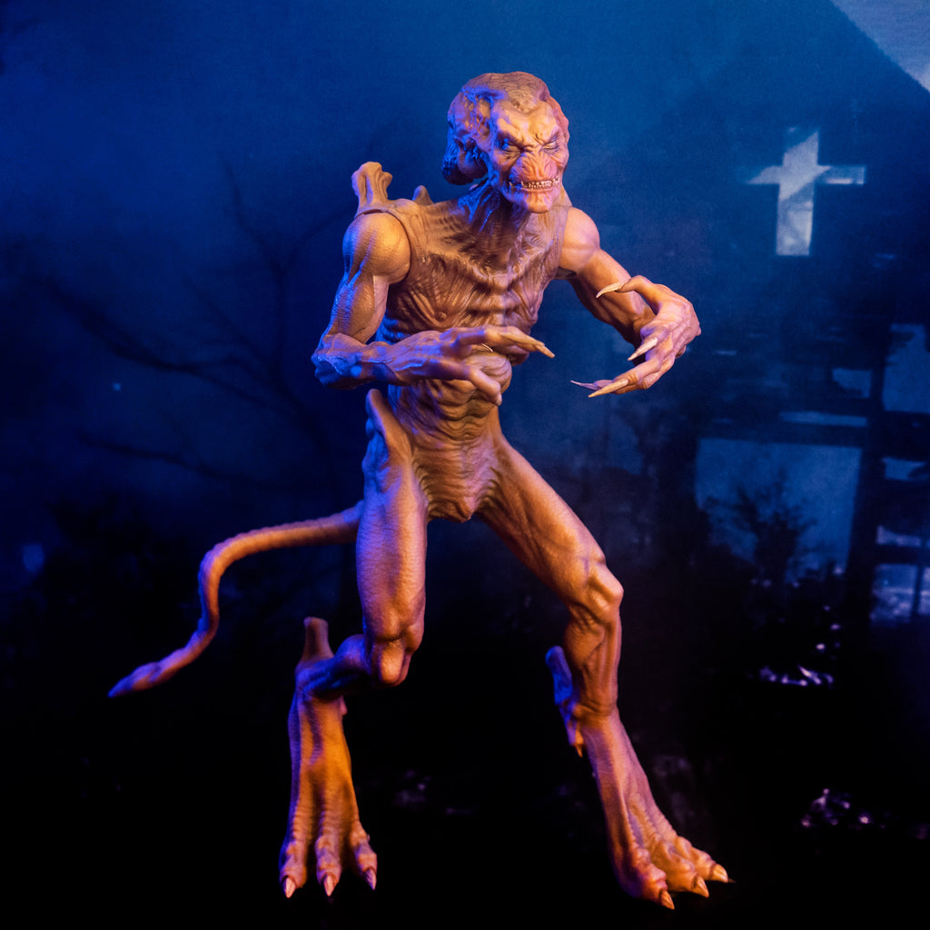 Misty blue and black glamour background, dilapidated church silhouette.  Slight right view Pumpkinhead 8" scale figure. Pale orange tan creature with enlarged upper head. White eyes, animal-like muzzle, mouth showing sharp yellow teeth. Bony with defined muscles, protrusions on shoulders, long limbs and tail. Hands have 4 long fingers and claws. Animal-like 3 toed feet with bony protrusions on heels, short claws.