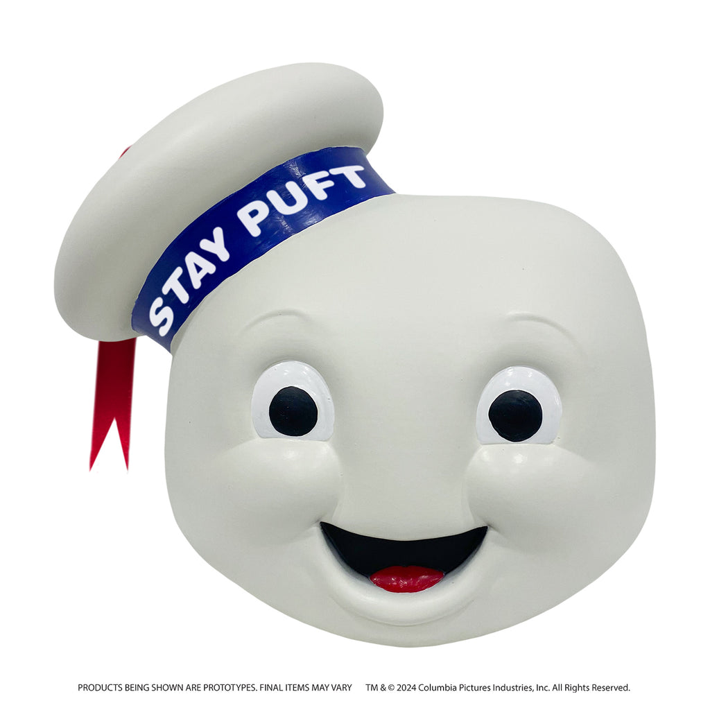 Mask, front view.  round white head, large eyes with large black pupils, mouth open in a smile showing red tongue.  White hat cocked to the side on the right side of the head, blue band with white text reads Stay Puft, red ribbon tassel hanging from the top center.