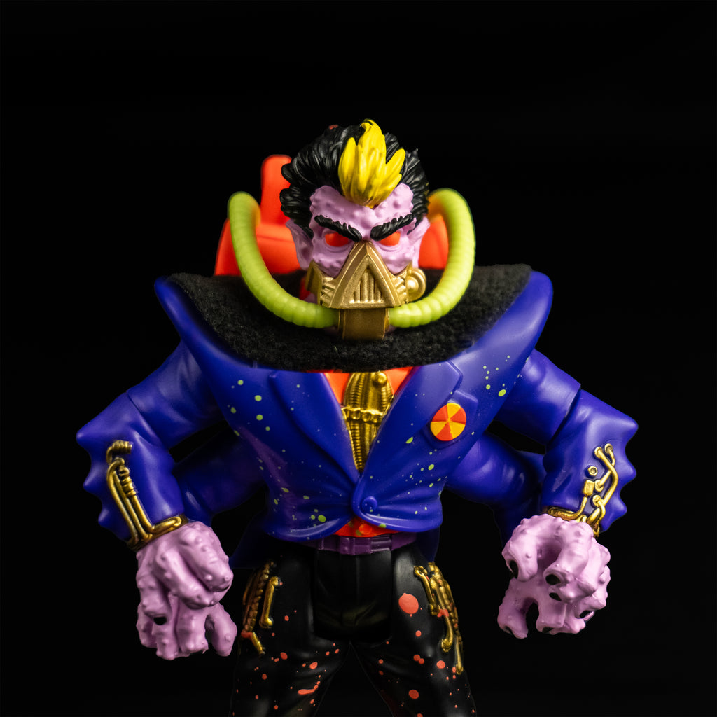 Black background. Action figure, front view closeup. Short black hair, yellow stripe in the center front. Pale purple bumpy flesh. Black bushy eyebrows, red eyes. gold mask, triangular over nose and mouth, horizontal bands around lower jaw, yellow hoses on sides attached to orange backpack. Wearing orange shirt with a gold tie, blue jacket, four arms, black cape, black pants with orange splatter.
