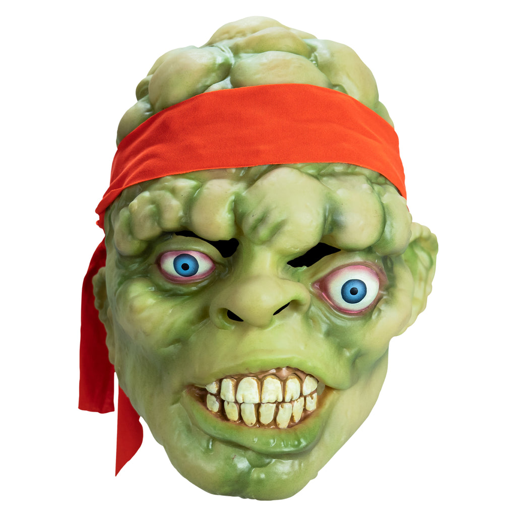 Mask, front view. Bald head, green lumpy flesh, with red-orange headband tied around forehead. Misaligned, red-rimmed blue eyes, crooked nose. Lips open showing large cracked teeth.
