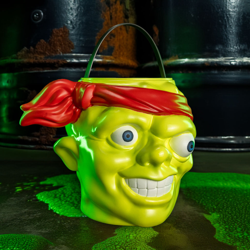 background with toxic waste barrels and green slime. Candy pail, slight right view.  Bald head, green lumpy flesh, with red-orange headband tied around forehead. Misaligned blue eyes, crooked nose. Lips open showing large teeth. Green handle at top.