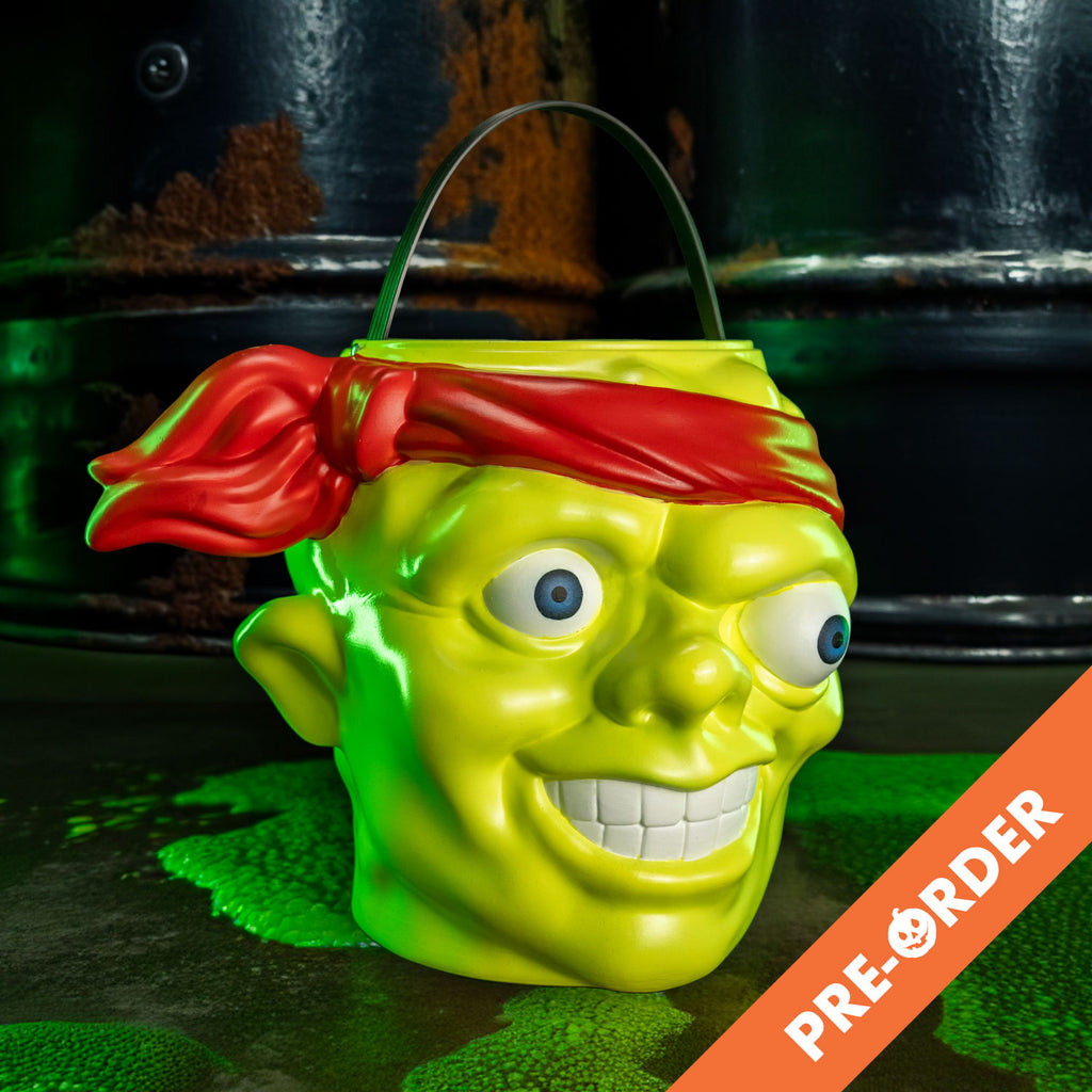 background with toxic waste barrels and green slime orange diagonal banner bottom right, white test reads pre-order.  . Candy pail, slight right view. Bald head, green lumpy flesh, with red-orange headband tied around forehead. Misaligned blue eyes, crooked nose. Lips open showing large teeth. Green handle at top.