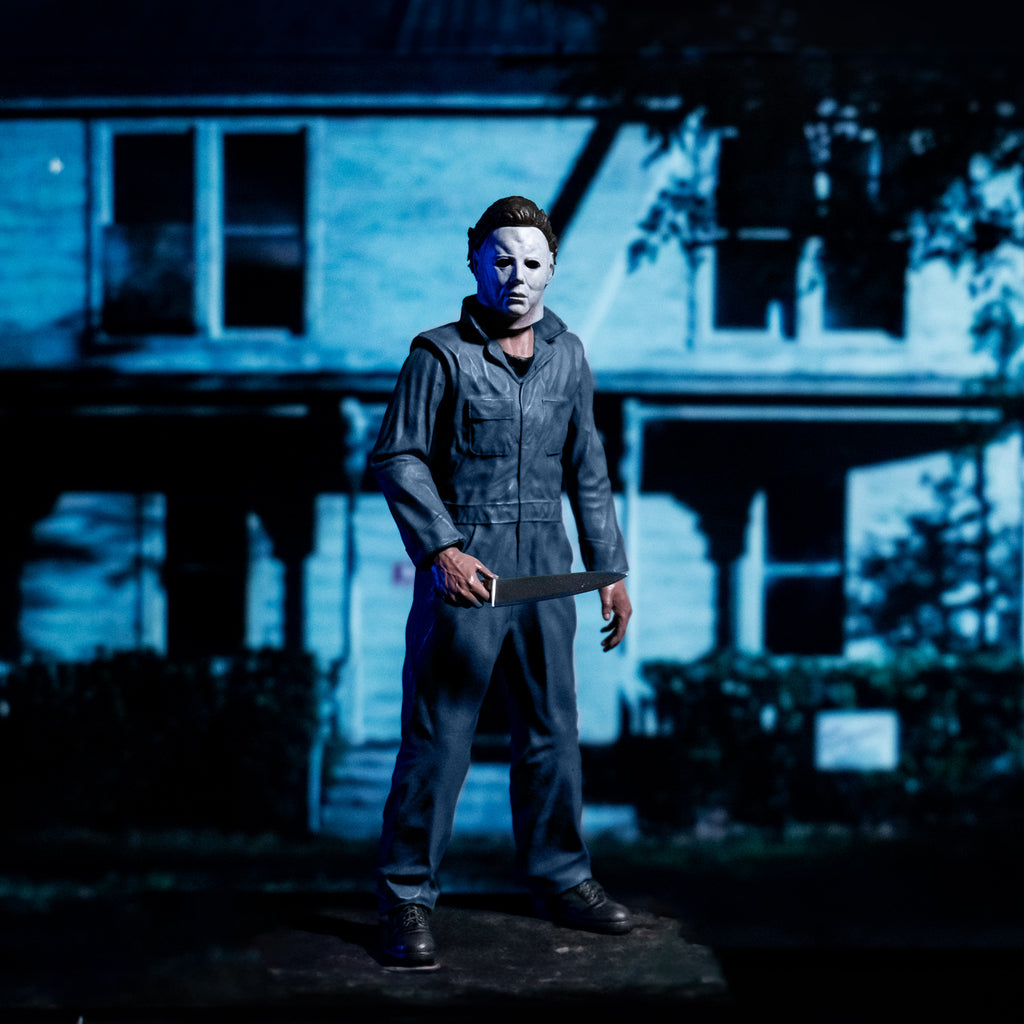 Glamour background, model house with nighttime lighting. Slight right full view in foreground Michael Myers 8 inch action figure. Wearing Halloween (1978) Michael Myers mask, dark coveralls, black boots. holding a kitchen knife in right hand. 