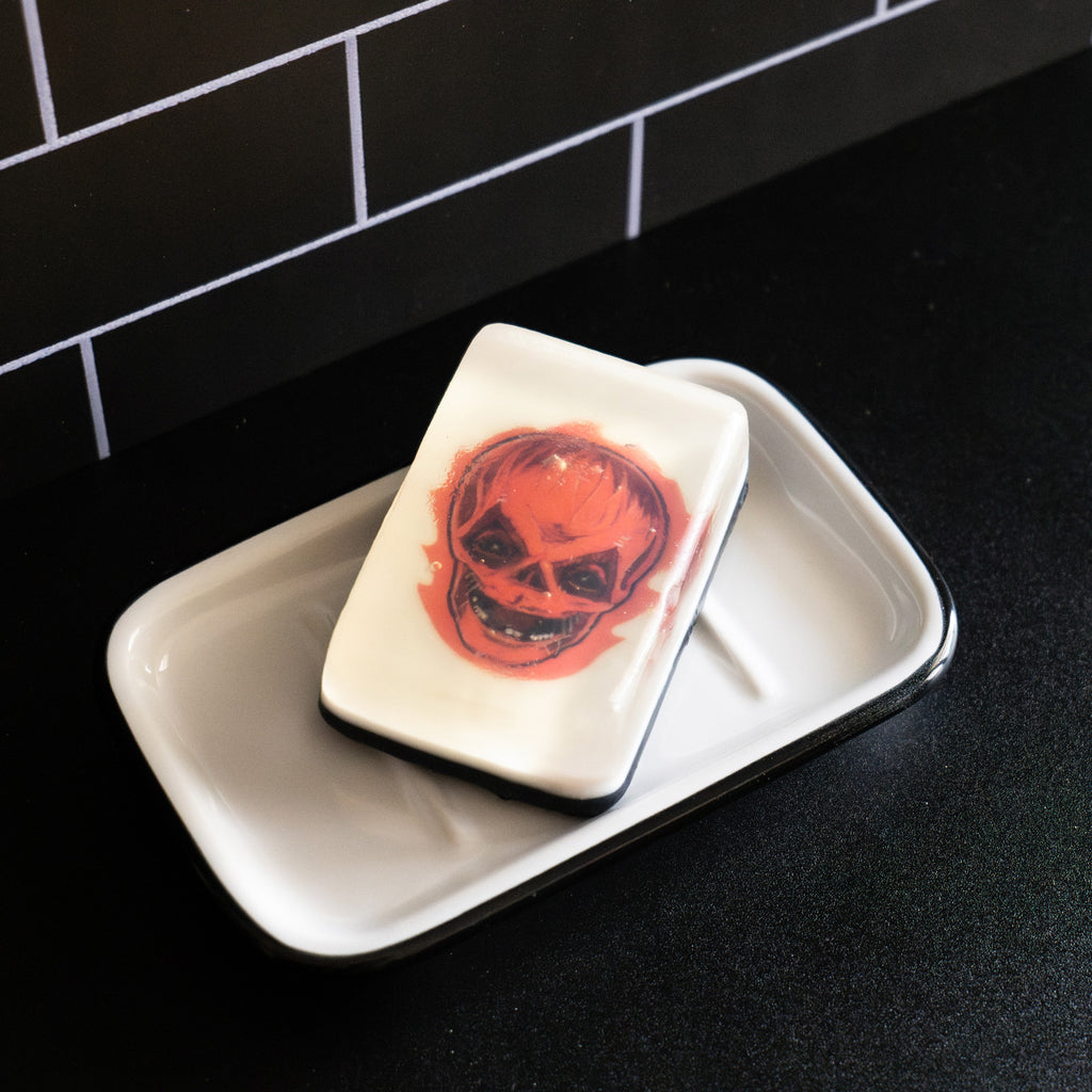Bar soap. White, with illustration of Trick 'r Treat Sam unmasked head under clear soap layer. Flaming orange jack o' lantern skull face. Shown in white soap dish on black counter.