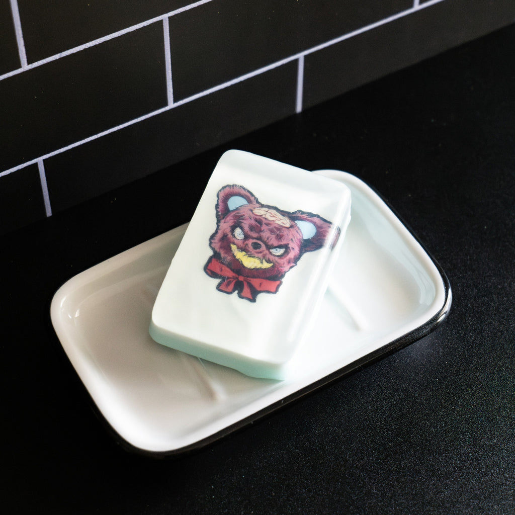 Bar soap. White, with illustration of Krampus Teddy Bear under clear soap layer. Brown teddy bear head, menacing grin with yellow teeth, brains exposed on scalp, red bow on neck. Shown in white soap dish on black counter.