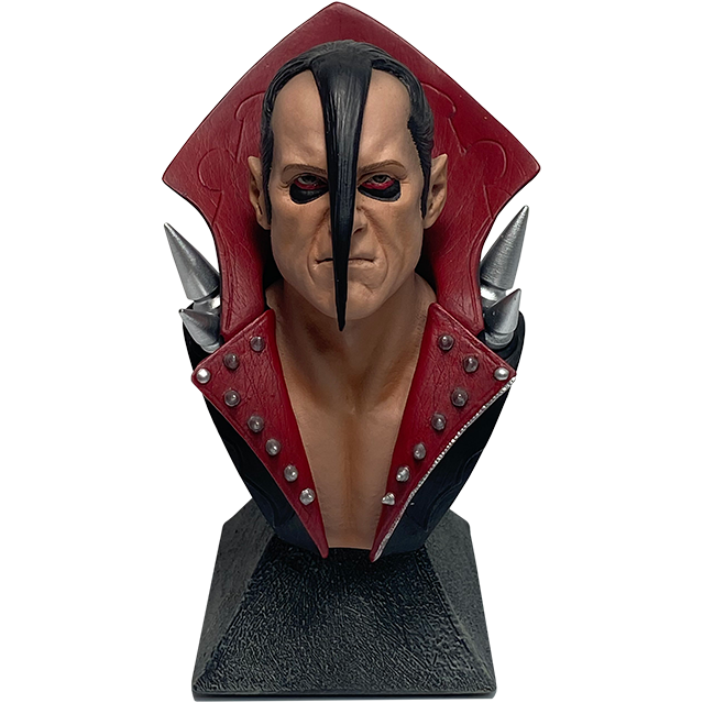 Mini bust. Head, shoulders and upper chest. Man with black and gray hair, with long lock pulled to the front and laid down the center of his face, wearing black coat with exaggerated tall red collar, large silver spikes on shoulders, smaller red collar at chest with small silver studs. Set on gray, stone textured base.