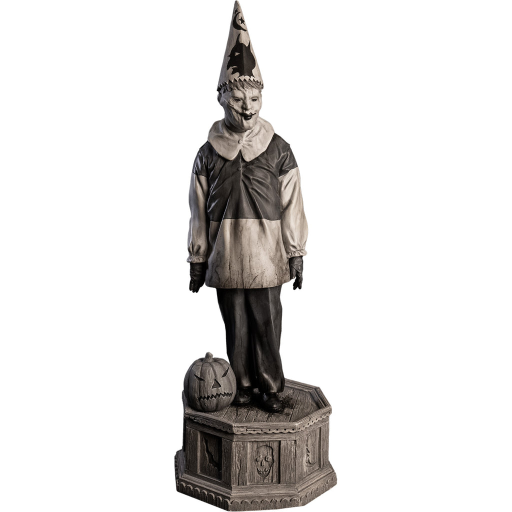 Front  view, statue. Grayscale, Creepy clown, tall pointy hat, black and white shirt with large white collar, black pants, shoes and gloves. Standing next to jack o' lantern on hexagon base made of gravestones
