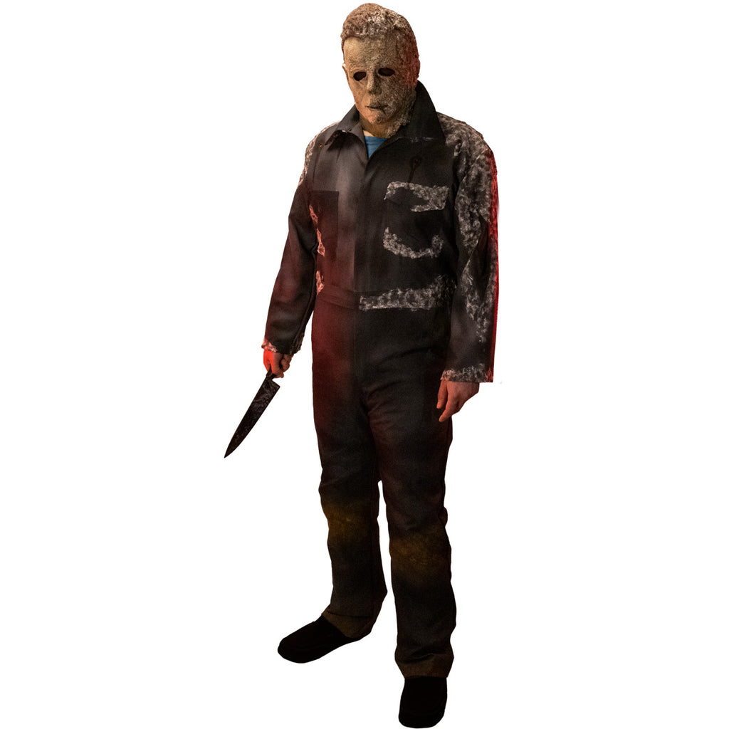 Man in Halloween Ends mask. Wearing dark coveralls, dirty distressed and appearing to be covered in mold. Holding knife in right hand.