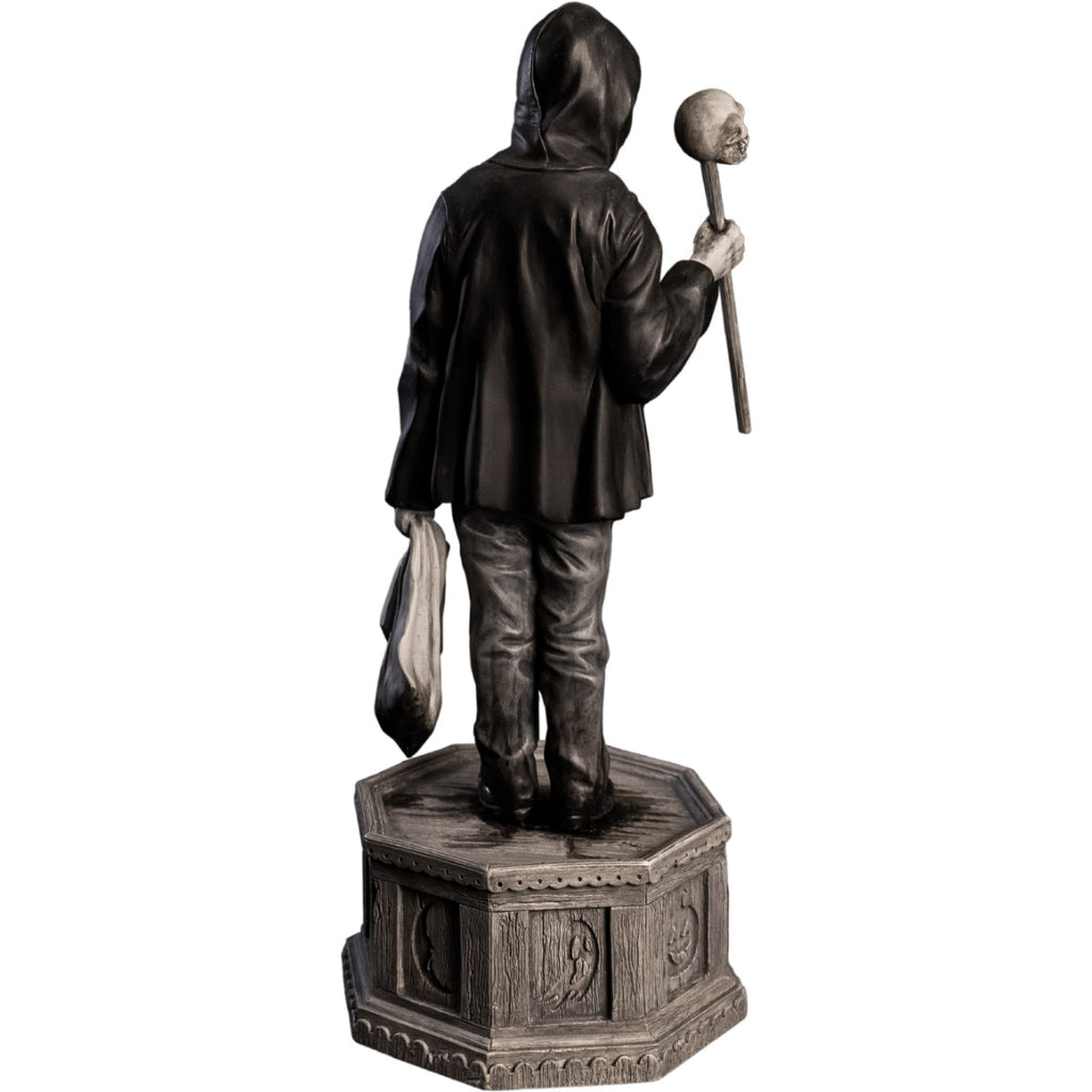 Back view, statue. Grayscale, Boy wearing black hooded jacket, jeans, black shoes. Holding baton topped with skull in right hand, holding dirty sack in left hand. Standing on hexagon base made of gravestones