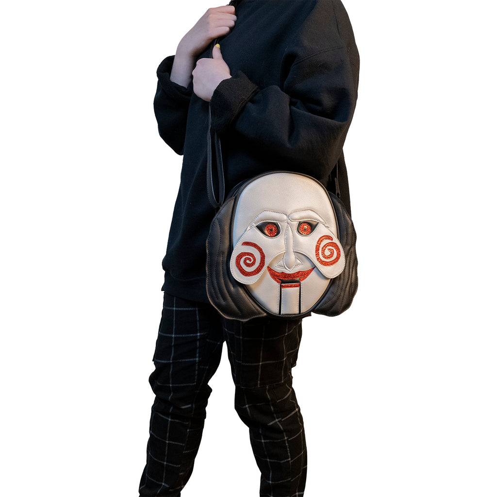 Person in black shirt and plaid pants, wearing bag, front view. Saw, Billy puppet face, black hair on sides, white face, black-rimmed red eyes, red spirals on cheeks, red lips on hinged ventriloquist dummy mouth. Black adjustable strap.