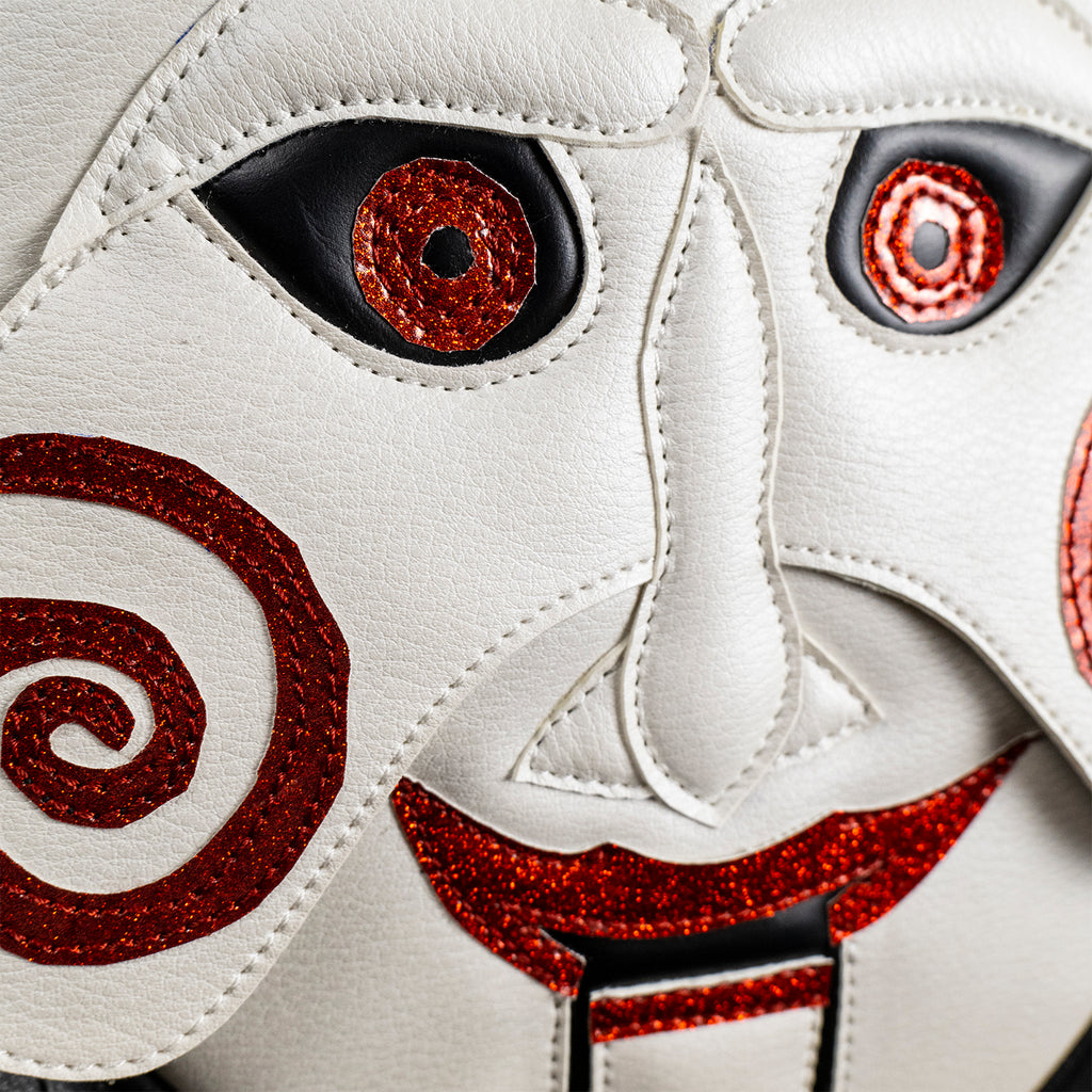 Close up of face on bag, front view. Saw, Billy puppet face, white face, black-rimmed red eyes, red spirals on cheeks, red lips on hinged ventriloquist dummy mouth. 