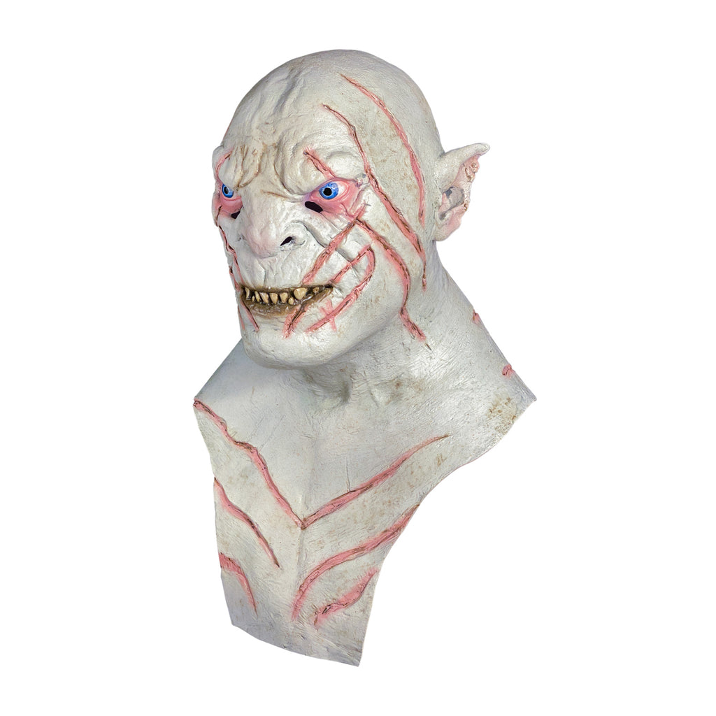 Mask, head, neck and chest, left side view. Orc, bald, white flesh, pointed ears, pink-rimmed blue eyes, sharp yellow teeth in mouth, dark pink stripes on head, face and chest.