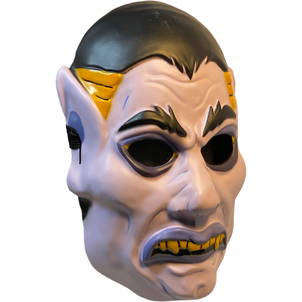 Plastic mask, right side view. White vampire face. Black hair, eyebrows and eyes. Blue-gray eyeliner and lips. Frowning mouth with yellow teeth and fangs. Yellow at temples. Pointed ears.