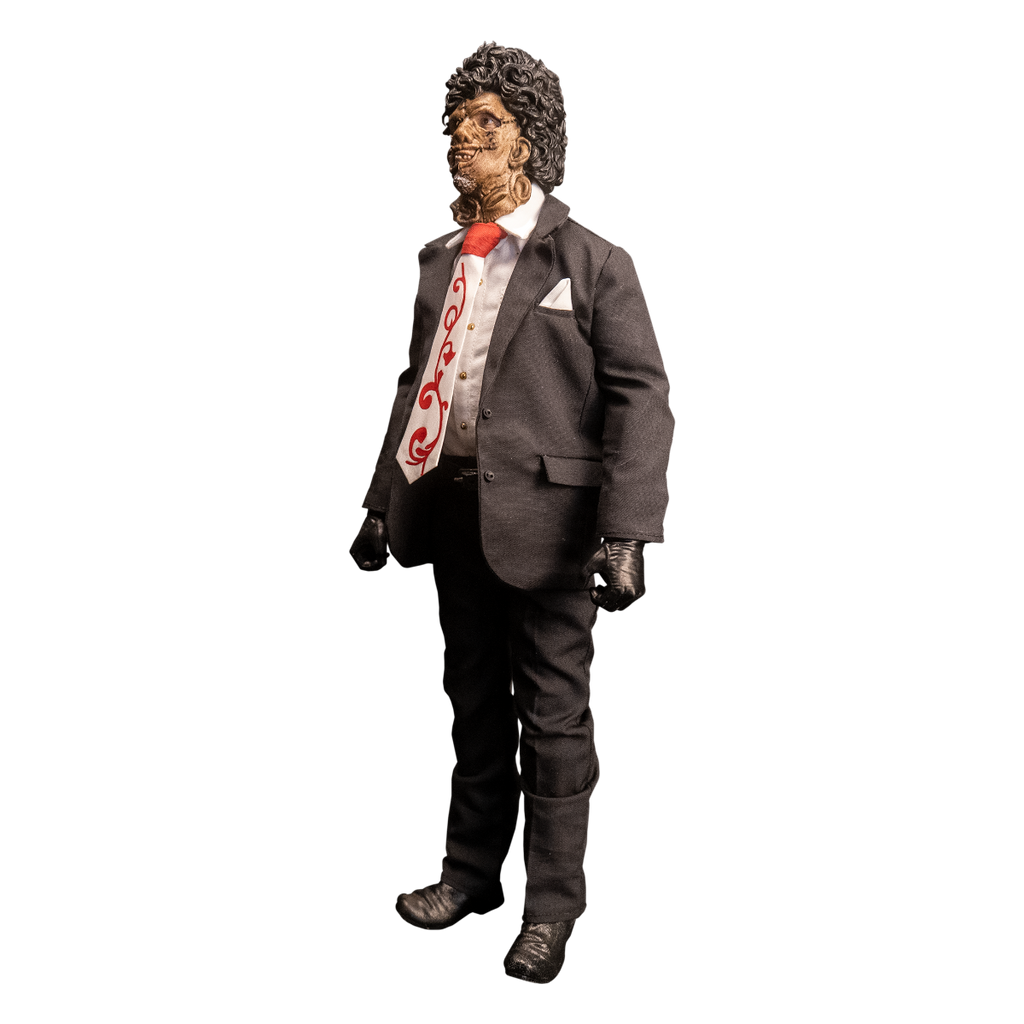 Action figure. Left view. Patchwork mask made of skin, brown hair. Wearing white collared shirt and orange and white necktie under dark suit coat, dark pants and belt. Black gloves and shoes.