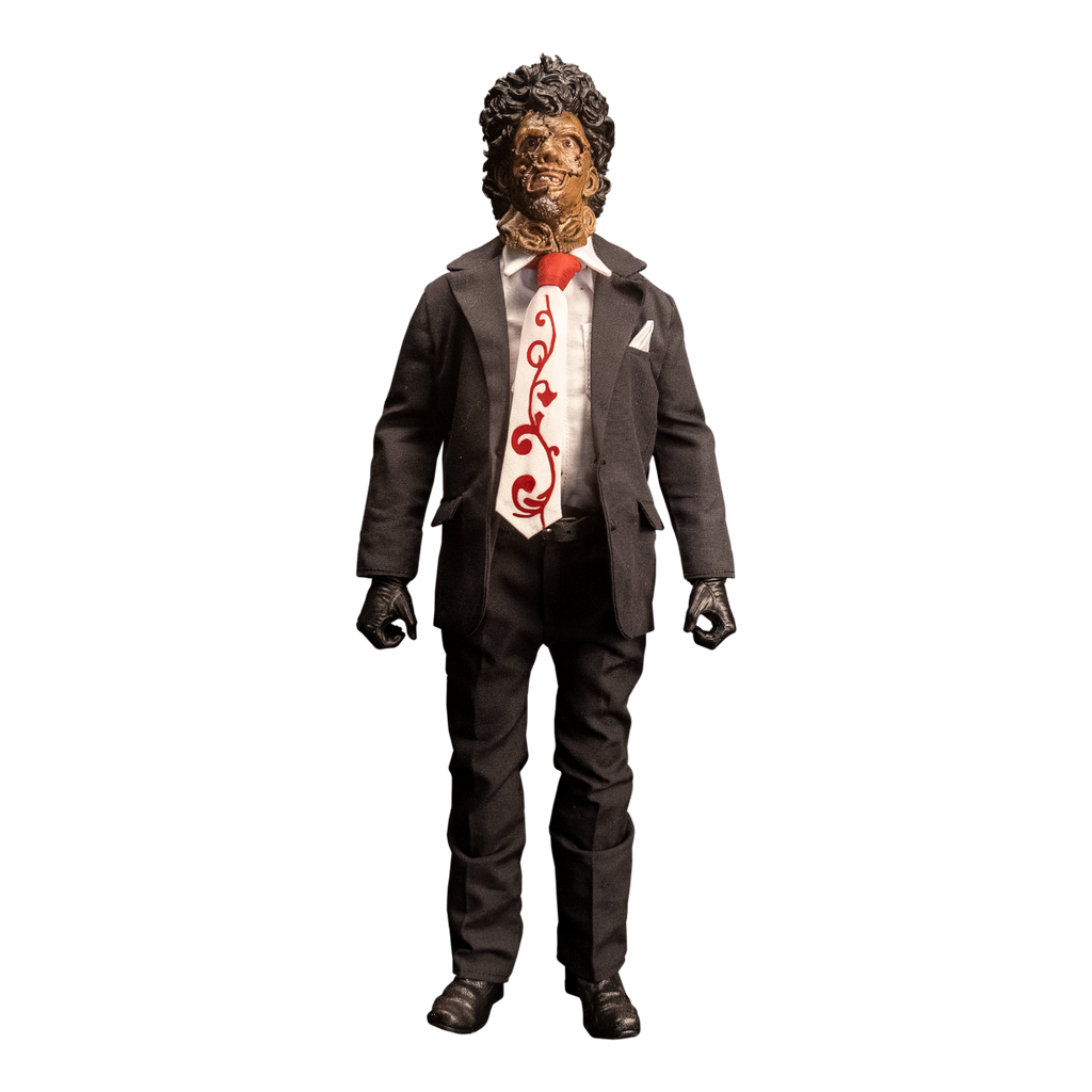 Action figure. Front view. Patchwork mask made of skin, brown hair. Wearing white collared shirt and orange and white necktie under dark suit coat, dark pants and belt. Black gloves and shoes.