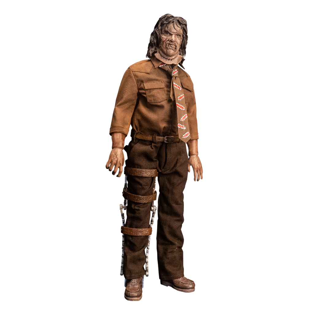 Action figure. Slight right view. Patchwork mask made of skin, brown hair. Wearing brown collared shirt and brown, red and white necktie, dark brown pants and belt. Brown shoes. Brown leg brace on right leg.