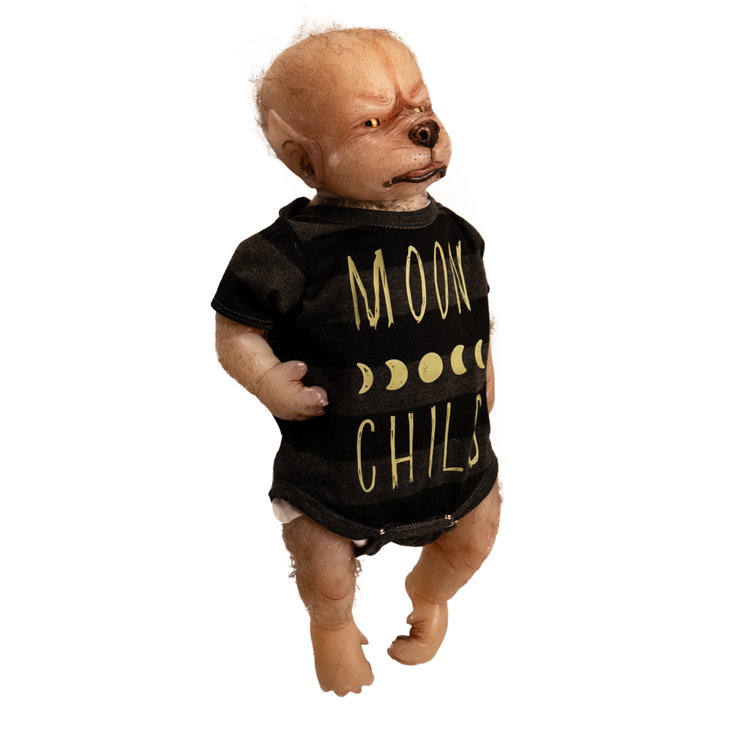 Doll. Full body right view, standing. Tan skin, pointed ears on side of head, sparse light fur on head and arms. Gold eyes, canine nose, mouth with pink tongue slightly sticking out. Paw like hands and feet with claws. Wearing black and gray striped short-sleeved onesie, yellow moon phases, Yellow text on shirt reads Moon Child
