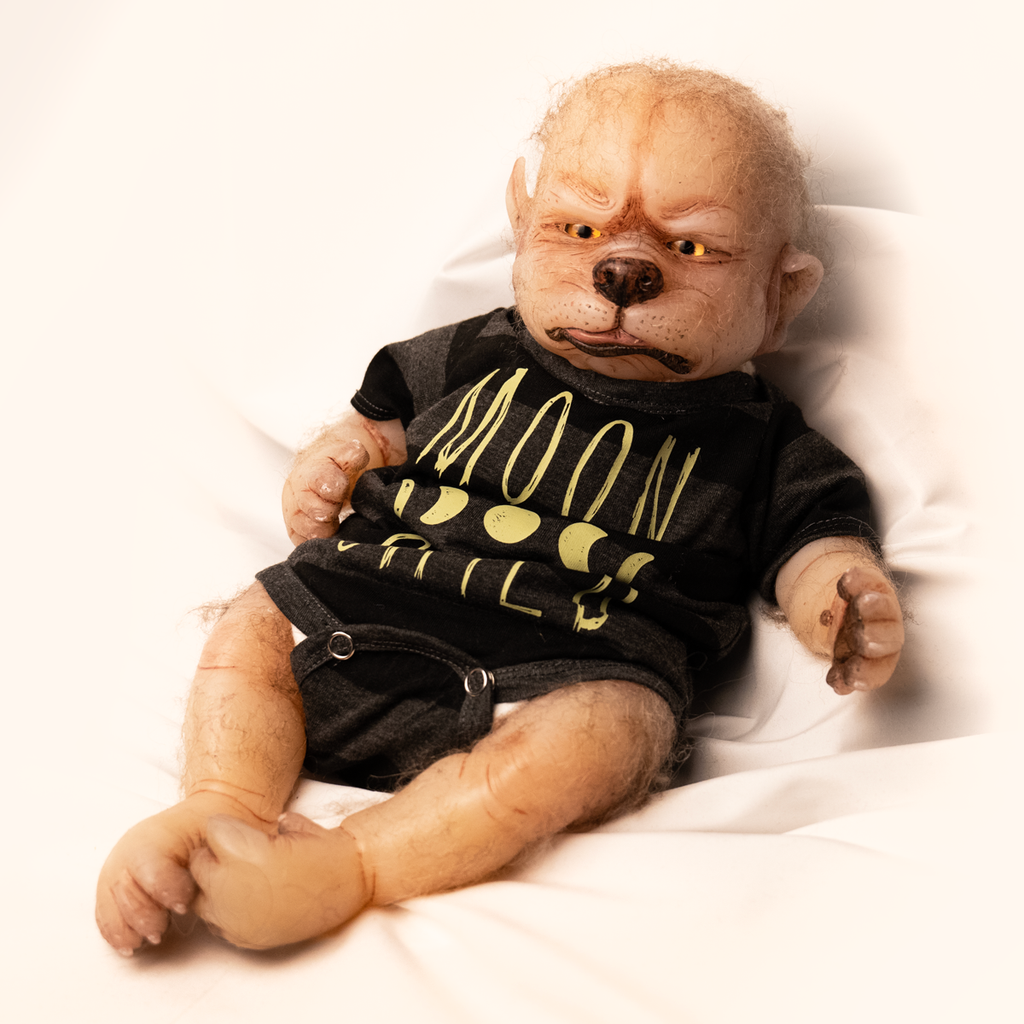 Doll. Full body view, reclining. Tan skin, pointed ears on side of head, sparse light fur on head and arms. Gold eyes, canine nose, mouth with pink tongue slightly sticking out. Paw like hands and feet with claws. Wearing black and gray striped short-sleeved onesie, yellow moon phases, Yellow text on shirt reads Moon Child