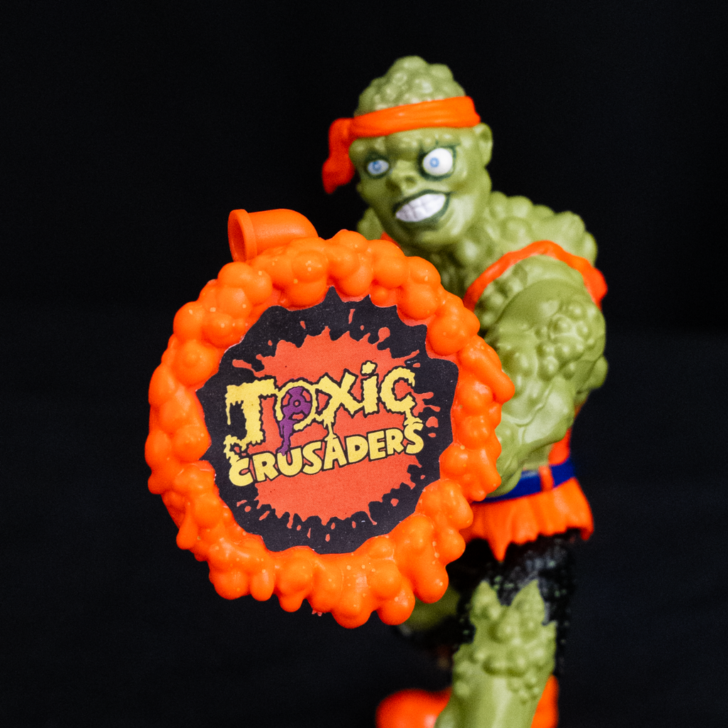 Toxie figure, black background, close up of shield accessory, lumpy orange shield, yellow text reads Toxic crusaders