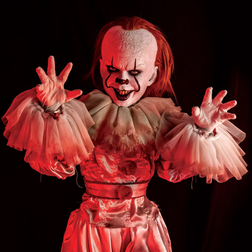 Pennywise doll, close up view under red lighting. clown face, red hair, white skin, large forehead, yellow eyes and nose, dark lips, creepy smile with crooked yellow buck teeth. White gloved hands outstretched, dirty neck ruffle, red trim on sleeves and belt, red pompoms.